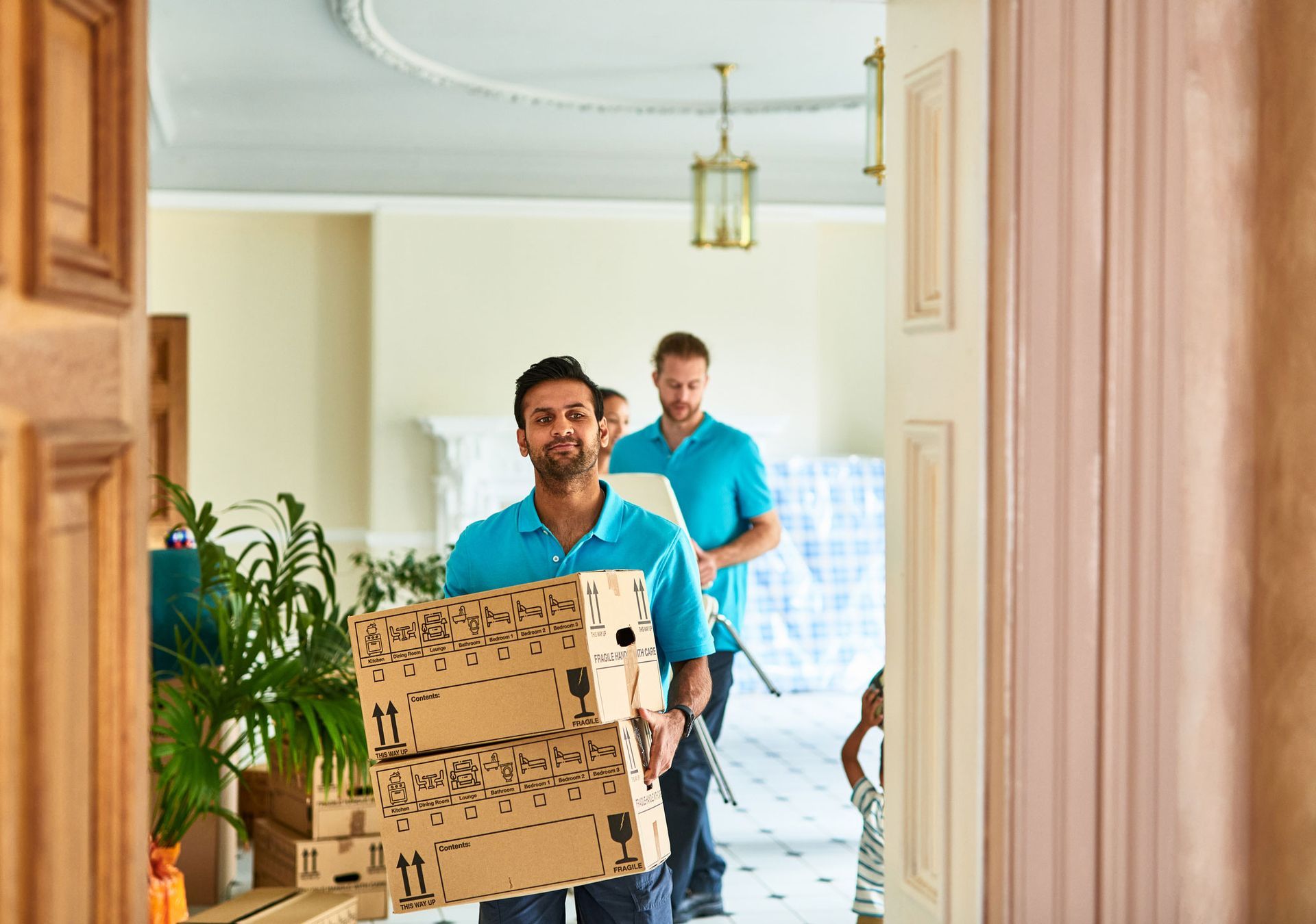 Two movers carry boxes into a house, with plants and furniture visible in the background setting Two movers carry boxes into a house, with plants and furniture visible in the background setting