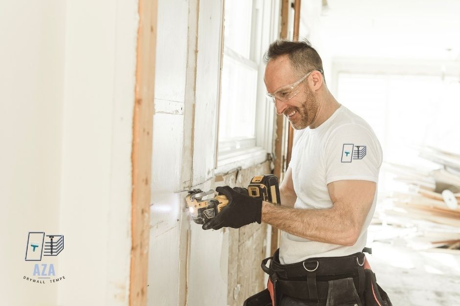 A drywall repair company professional in a white t-shirt uses a circular saw to cut through drywall in an unfinished residential room with exposed studs and bright natural lighting from windows near E Amberwood Dr, and S 44th Way, Ahwatukee Foothills, AZ.