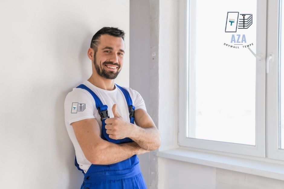 A drywall repair company contractor stands with arms crossed, wearing blue overalls and a white t-shirt. He is smiling in a bright, modern residential construction site with white walls and large windows, ready to provide professional services near S Palo Verde Dr, and W Seaver Ave, Apache Junction, AZ.