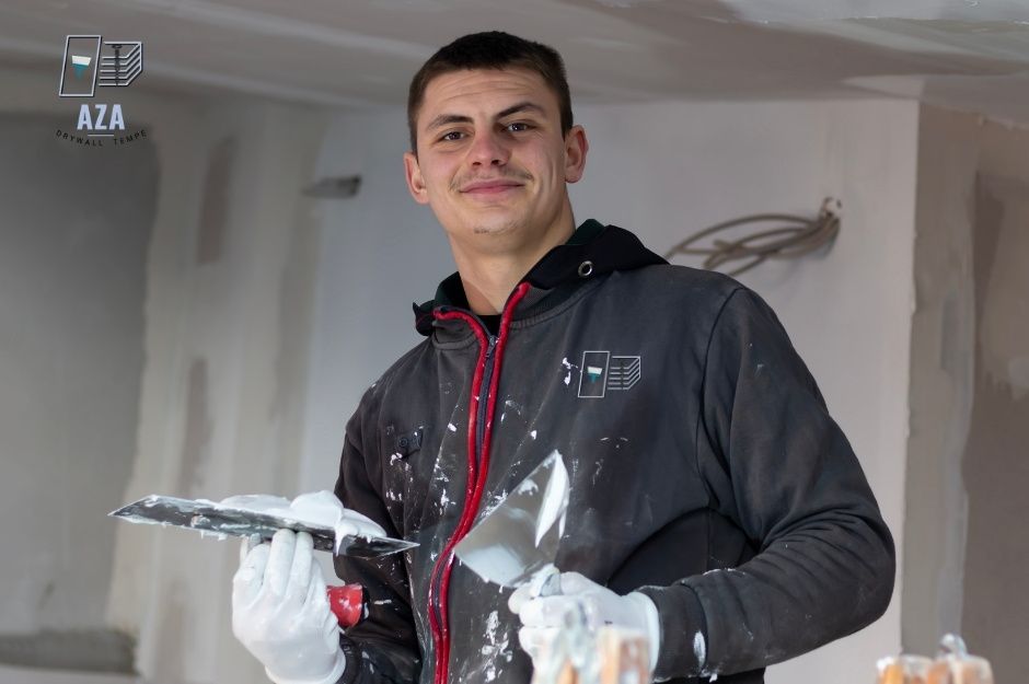 A drywall repair company worker in dark hoodie holds a putty knife for residential construction with joint compound while standing in a white-walled room near N Spur Cross Rd, and Maguay Rd, Cave Creek, AZ.