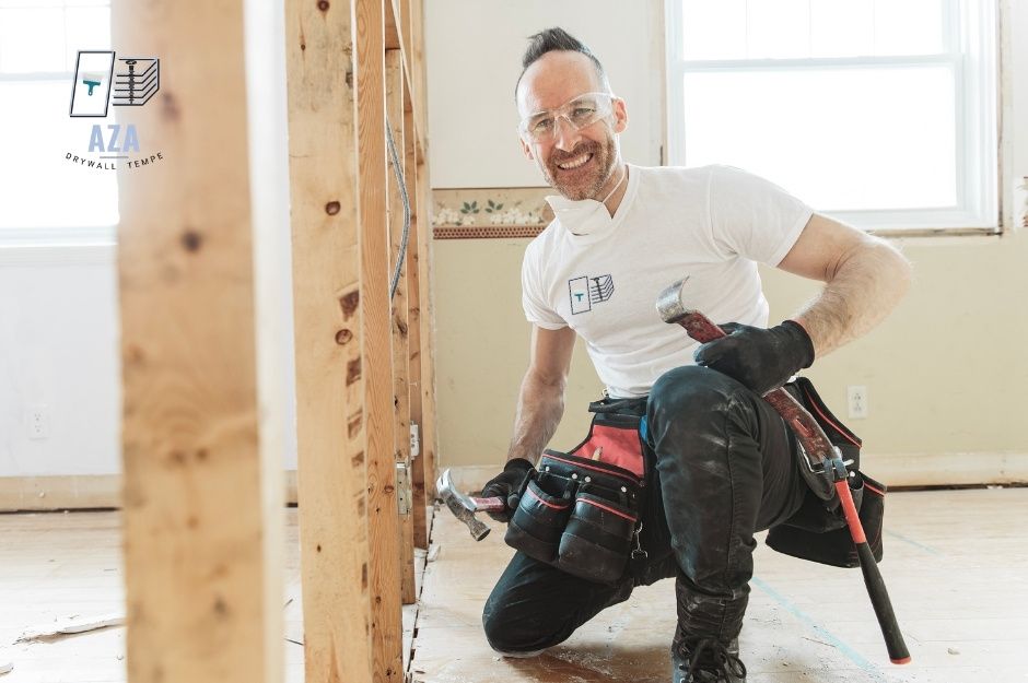 A drywall repair company professional kneels while working on wooden wall framing. The smiling worker wears a white t-shirt and dark work pants, holding a hammer. Wood studs are visible in the background of the residential construction site near S Ellesmere St, and E Wimpole Ave, Gilbert, AZ.