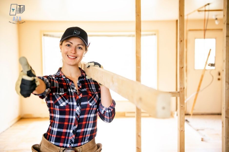 A drywall repair company worker wearing a red and black plaid shirt, holding a piece of wood while giving a thumbs-up gesture. This smiling female technician is in an interior residential construction space with wooden framing visible. The project is near S Thoroughbred Ln, and E E Wallaby Way, Mayer, AZ.