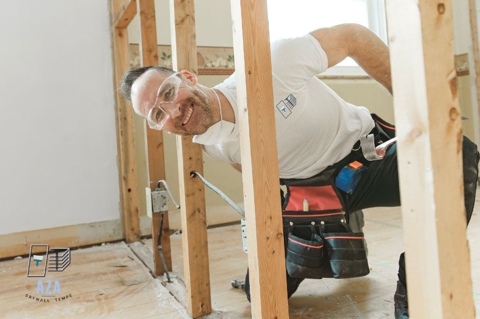 A drywall repair company worker kneels between wooden wall studs during residential construction work. The person wears work clothes and appears to be examining or working on the framing. Construction tools and materials are visible in the background near E Desert Park Pl, and N 47th St, Paradise Valley, AZ.
