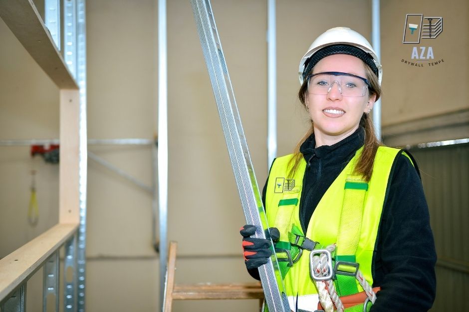 A drywall company worker smiling while holding an aluminum drywall panel in an unfinished room with exposed metal framing and drywall sheets. She wears a yellow safety vest, a white hard hat, and safety glasses on a residential construction site near N 41st Dr, and W Granite Basin Dr, Rio Vista, AZ.