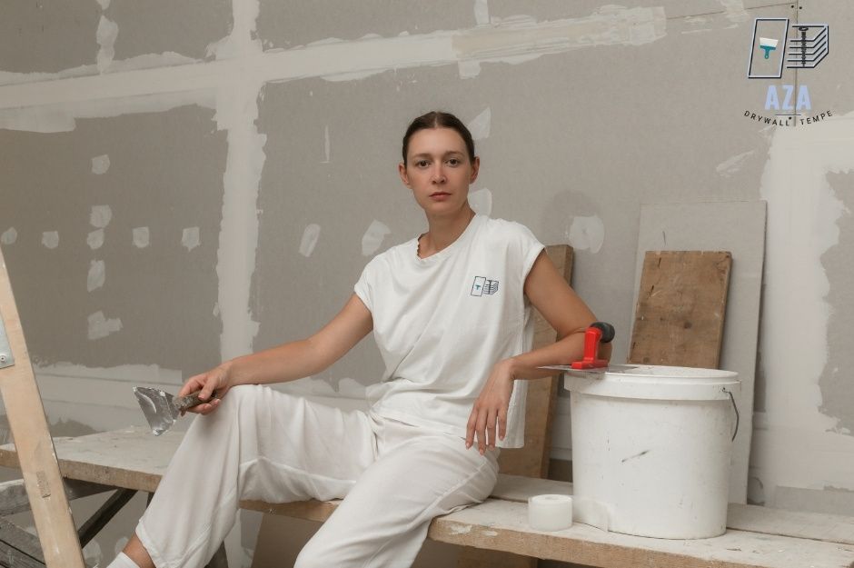 A drywall repair company worker at a residential construction site sits on wooden planks holding a putty knife, with a paint bucket and roller nearby in an unfinished room near S Desert Burst Ct, and E Cherrywood Dr, Sun Lakes, AZ.