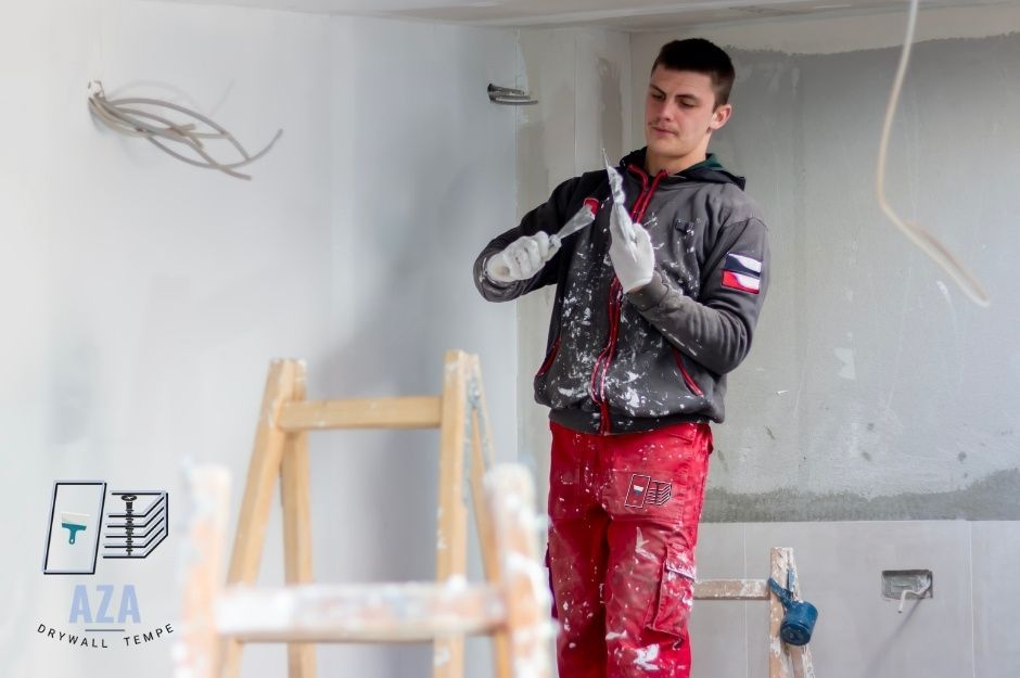 A drywall repair contractor at a commercial construction site wearing a black hoodie and red work pants, using a drywall knife to apply joint compound to the white wall, with a wooden ladder visible near N 52nd Ave, and W Tonopah Dr, Glendale, AZ.