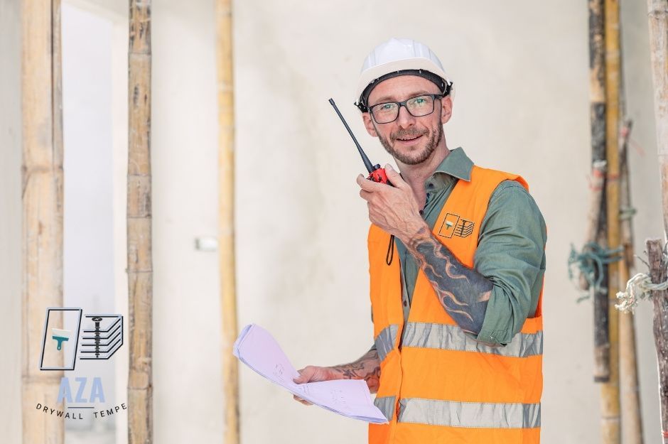 A drywall repair contractor is wearing a white hard hat, glasses, an orange high-visibility vest over a green shirt, holding a walkie-talkie radio and blueprints in a commercial construction site with exposed wall framing near W Black Hill Rd, and N 92nd Dr, Peoria, Arizona.