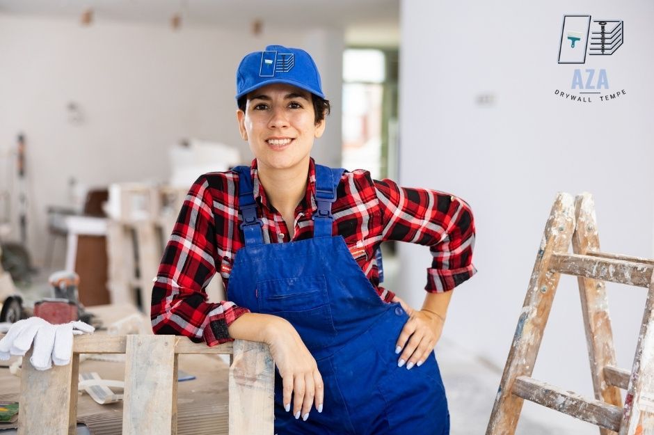 A drywall repair contractor smiles while leaning on a wooden sawhorse in a commercial construction site workspace. She wears blue overalls, a red plaid shirt, and a blue cap. A wooden stepladder and work tools are visible nearby in the bright room near N 88th Ave, and W Adams St, Tolleson, AZ.