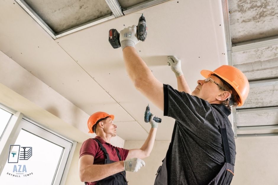 Drywall repair contractors work on ceiling installation. Two workers in orange hard hats and work clothes use cordless drills to secure drywall panels to the metal ceiling framework inside a residential construction site near W Hopi St, and Jessie May Ave, Avondale, AZ.