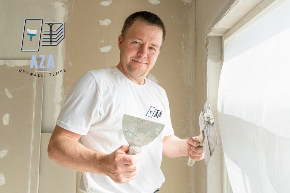 A drywall repair handyman wearing a white T-shirt stands near a window, holding a metal putty knife and a joint knife covered with joint compound while repairing drywall seams in a residential room with beige walls and white patches. Located near W Court St, and North Point Ridge Rd, Buckeye, AZ.