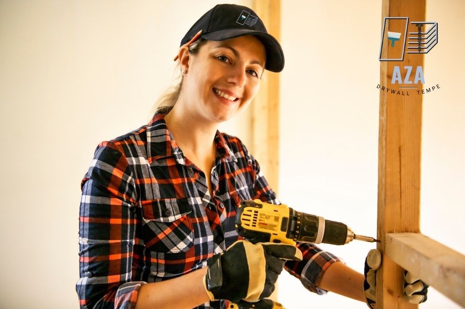 A drywall repair handyman in a black cap and plaid shirt smiles while holding a power drill, working on plasterboard repair at a residential construction site in a room with white walls and wood trim near E Palo Verde Dr, and Val Vista Dr, Cordes Lakes, AZ.