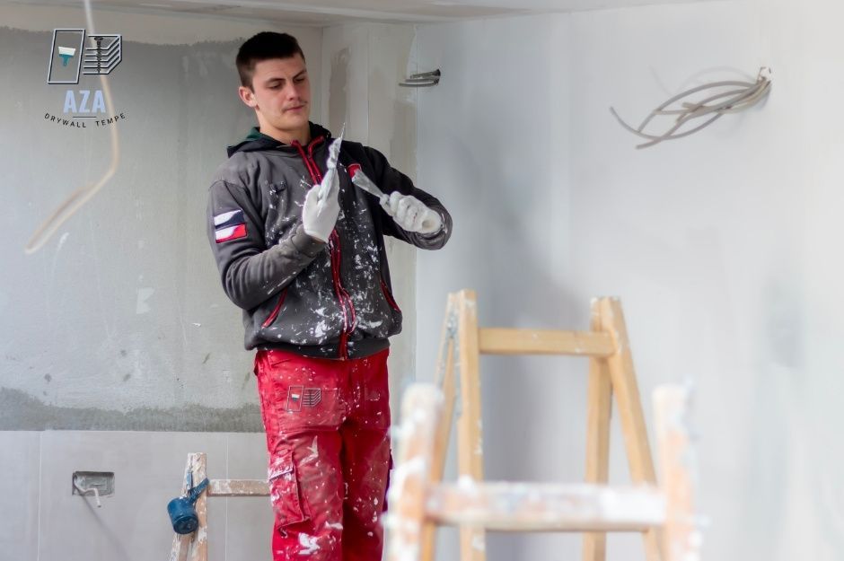 A drywall repair specialist wearing a gray jacket and jeans is holding a puffy knife while working on a residential construction site with exposed ceiling and a wooden ladder in the background near E Paint Pony, and Paint Pony Dr, Carefree, AZ.