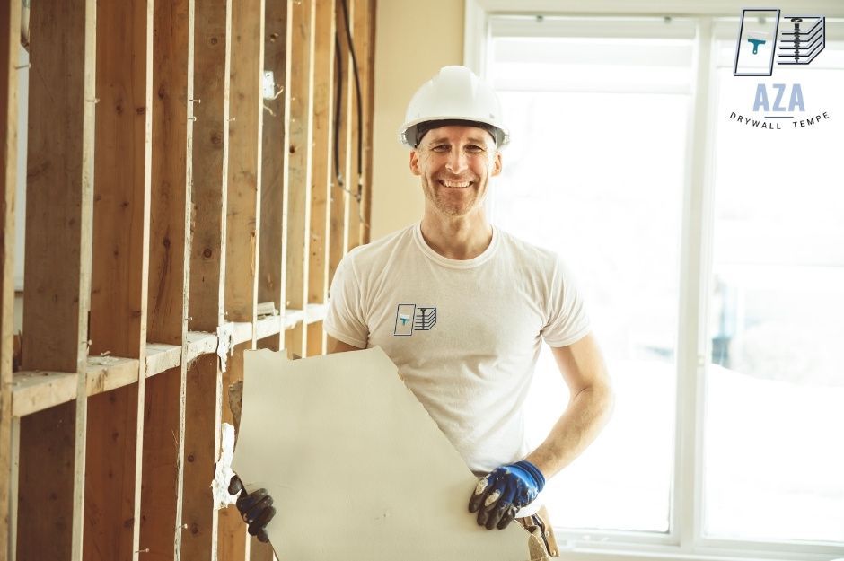A drywall repair specialist wearing a white hard hat and t-shirt smiles while holding a large sheet of drywall in a residential construction site. The worker wears blue work gloves and stands among exposed wooden wall studs with natural light from windows near W Harding Ave, and S 3rd St, Coolidge, AZ.