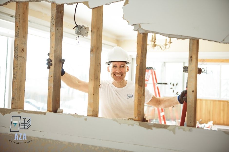 A drywall repair specialist in a white hard hat smiles while holding a sledgehammer during an interior renovation project, standing among exposed wooden ceiling joists and damaged drywall in a residential space. Located near W Cactus Wren Way, and N Monument Dr, Florence, AZ.