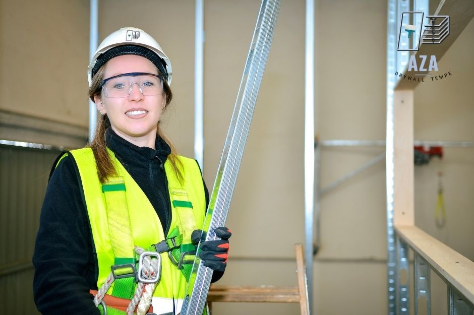A drywall repair specialist is smiling while holding an aluminum drywall panel in a framed interior space with exposed metal studs and drywall sheets. She wears a yellow safety vest, white hard hat, and safety glasses at the residential construction site near S Liberty School Rd, and W MC 85, Liberty, AZ.