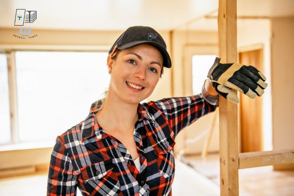 A drywall repair technician smiling while wearing a plaid shirt, cap, and protective gloves. She is leaning against a wooden frame inside a bright, unfinished room in a residential construction site near E Eagle Dr and E White Dr, Dewey–Humboldt, AZ.