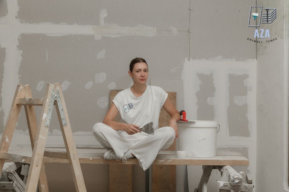 A drywall repair technician sits cross-legged on wooden sawhorses at a commercial construction site, holding a putty knife while taking a break from patching work. She wears white work clothes, surrounded by ladders and joint compound buckets near N Valley Rd, and W Shedd Rd, Eloy, AZ.