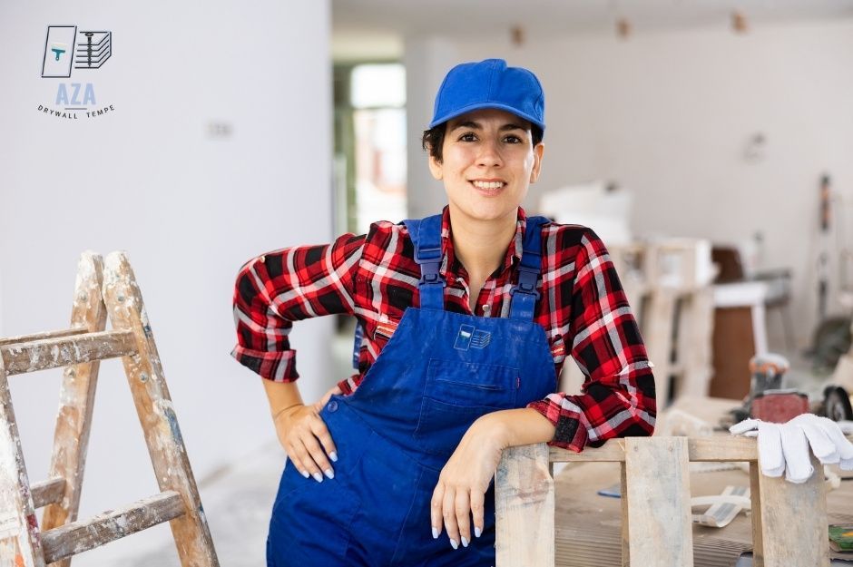 A drywall repair technician in blue overalls and red plaid shirt smiles in a residential construction workspace. She wears a blue cap and stands near a wooden ladder with white walls and renovation materials visible in the background at E Sabrosa Dr, New River, AZ.