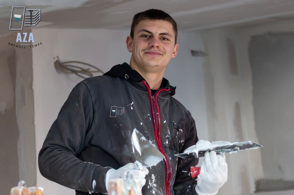A drywall repair technician in a dark hoodie holds a putty knife and trowel with white compound in an unfinished residential project near W Camino Del Sol, and W Sandridge Dr, Sun City West, AZ.