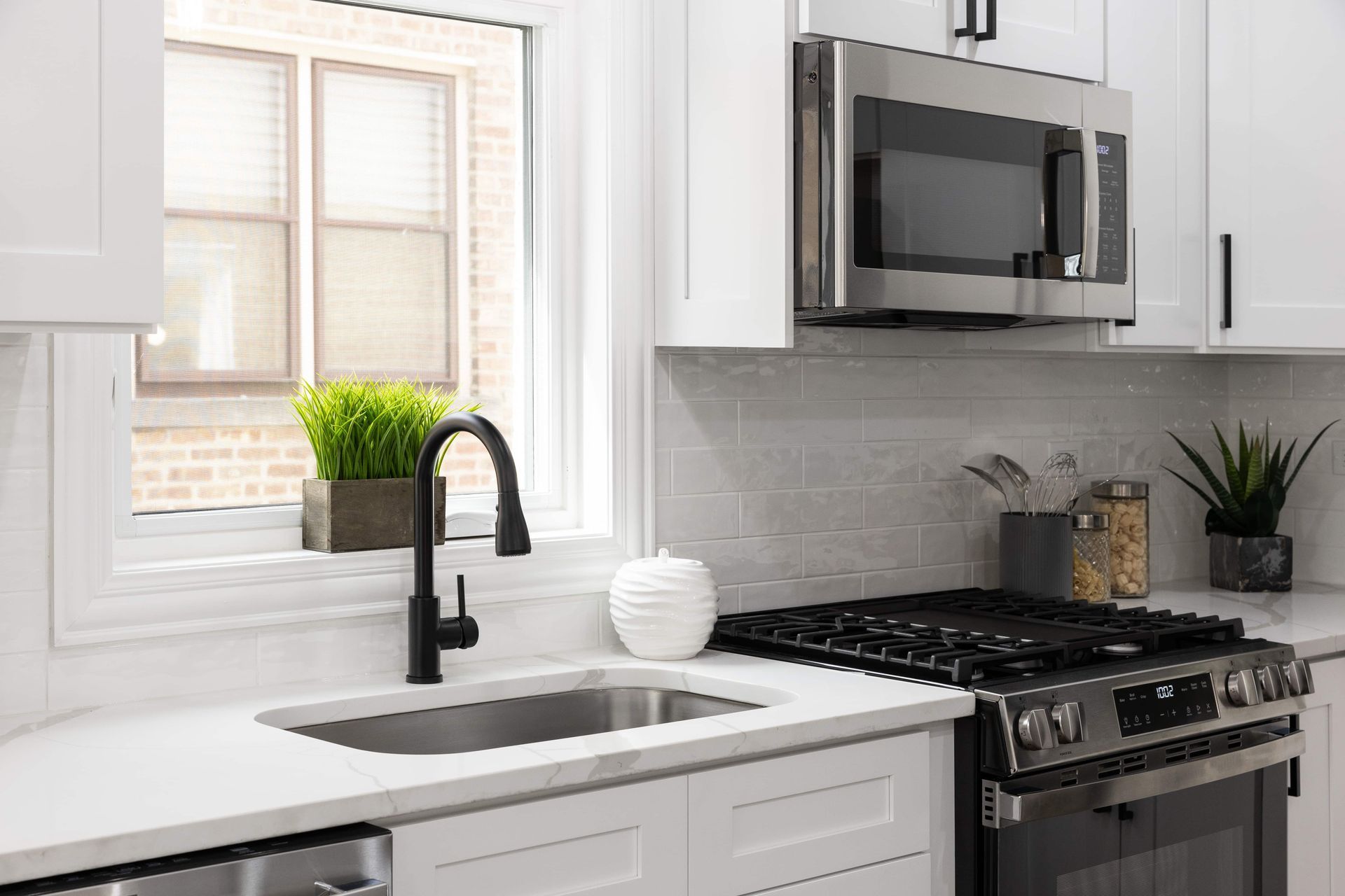 Modern kitchen with white cabinets, stainless steel appliances, and a black faucet. A window with a plant is over the sink.