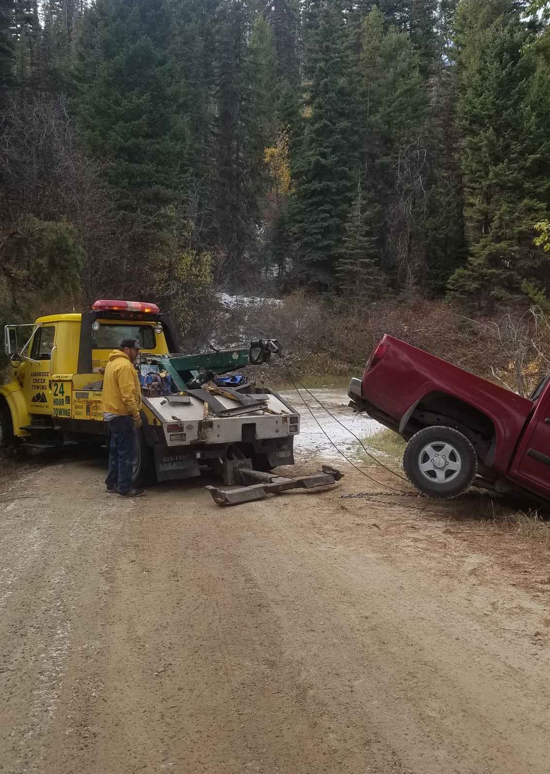 A tow truck pulling a red pickup truck out of a ditch on a dirt road, near a forest.