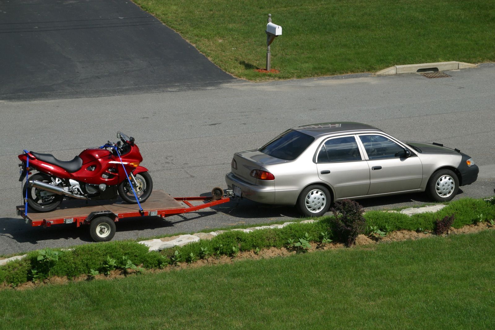 Red motorcycle on a trailer hitched to a silver car, parked on asphalt near a grassy lawn.