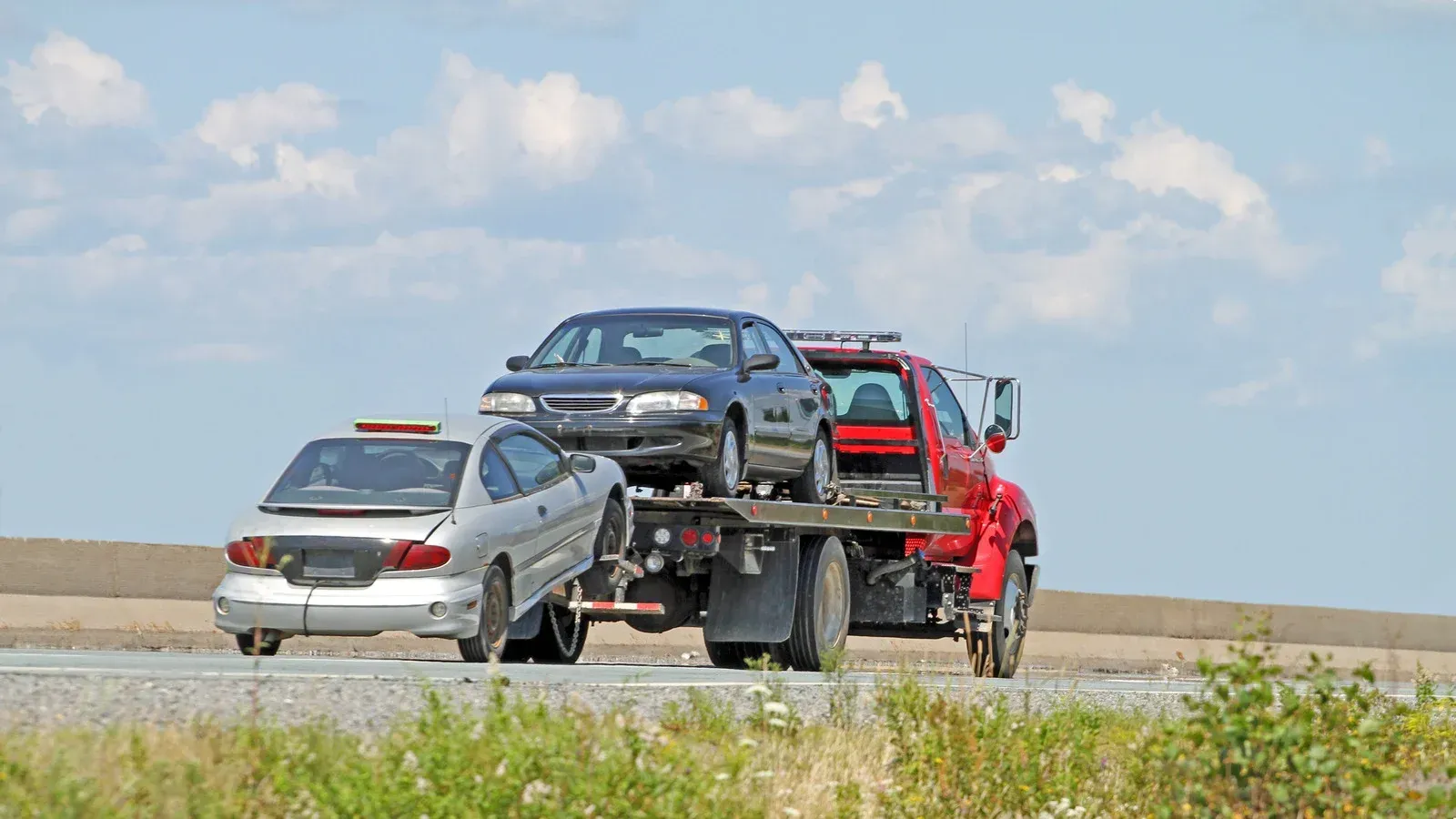 Tow truck hauling two damaged cars on a highway under a partly cloudy sky.