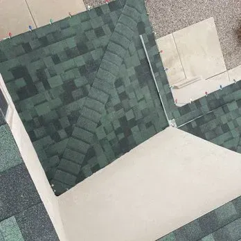 Green shingled roof with a corner, showing the edge of the roof and a concrete surface.