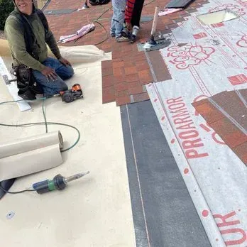 Roofer kneeling, installing shingles; rolls of material, tools on roof.