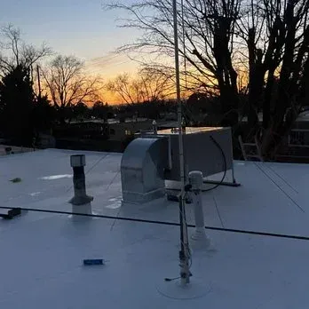 Rooftop with equipment, antenna, and sunset in the background. White flat roof with trees and buildings.