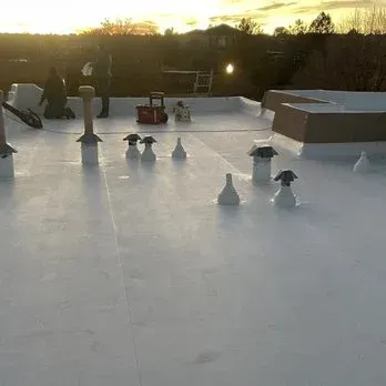 Flat white commercial roof with vents and skylight. Workers in the background. Evening sunlight.