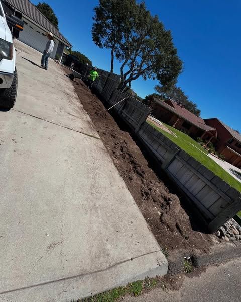 Sloped driveway with dirt and a leaning fence. A person works on the edge. Sunny day.