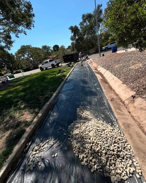 Workers laying gravel on black landscaping fabric in a roadside area.