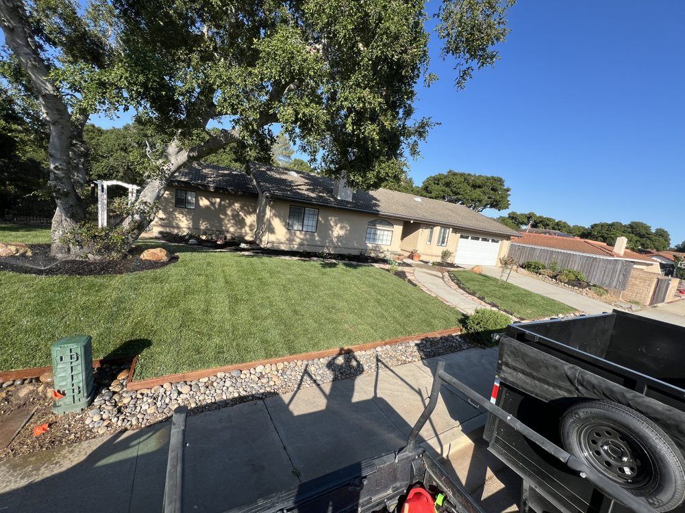 House with fresh green lawn, tree, and gravel landscaping. Trailer in foreground. Sunny day.