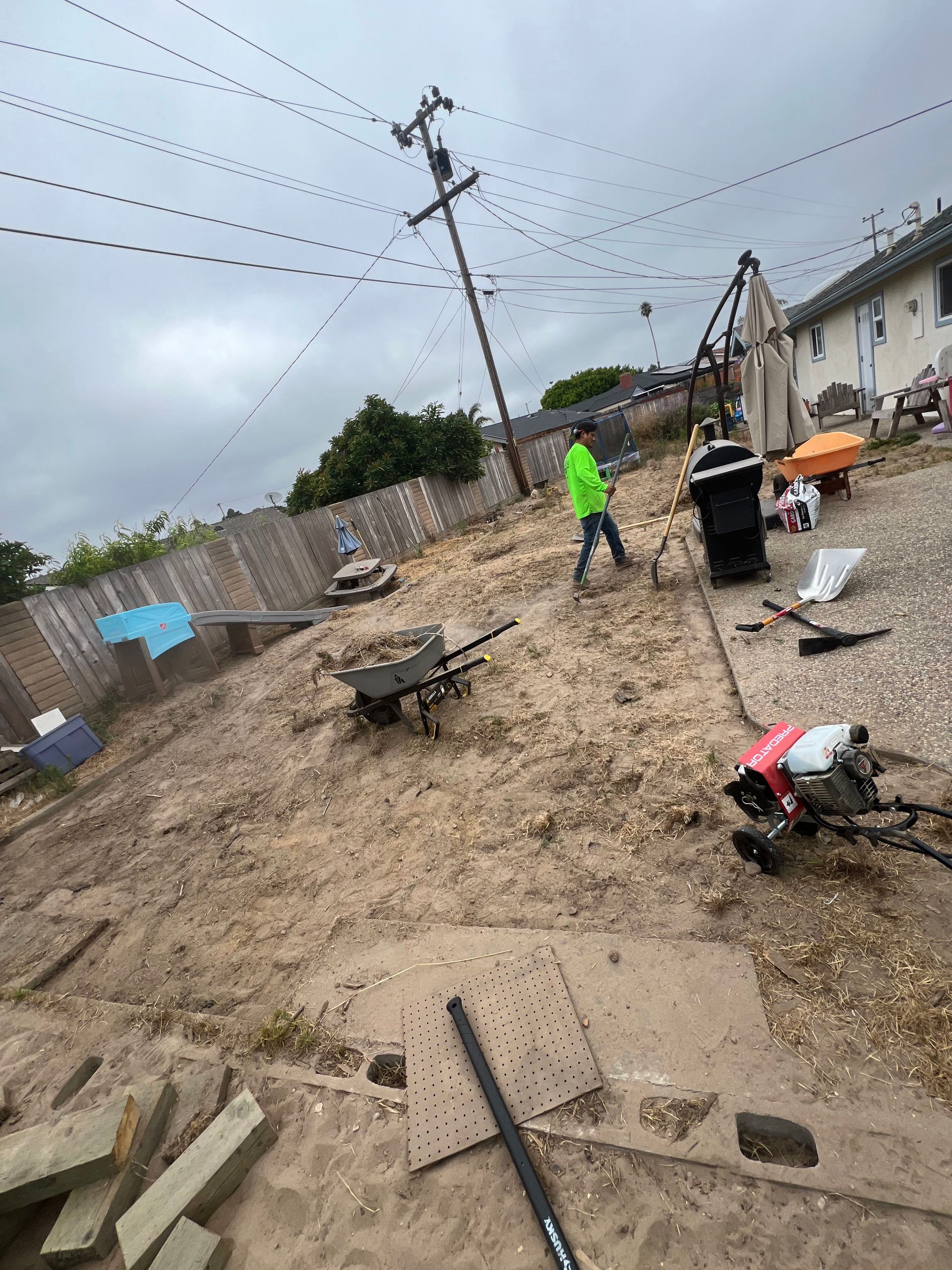 A person in a green jacket works in a yard, soil and construction equipment visible, overcast sky.
