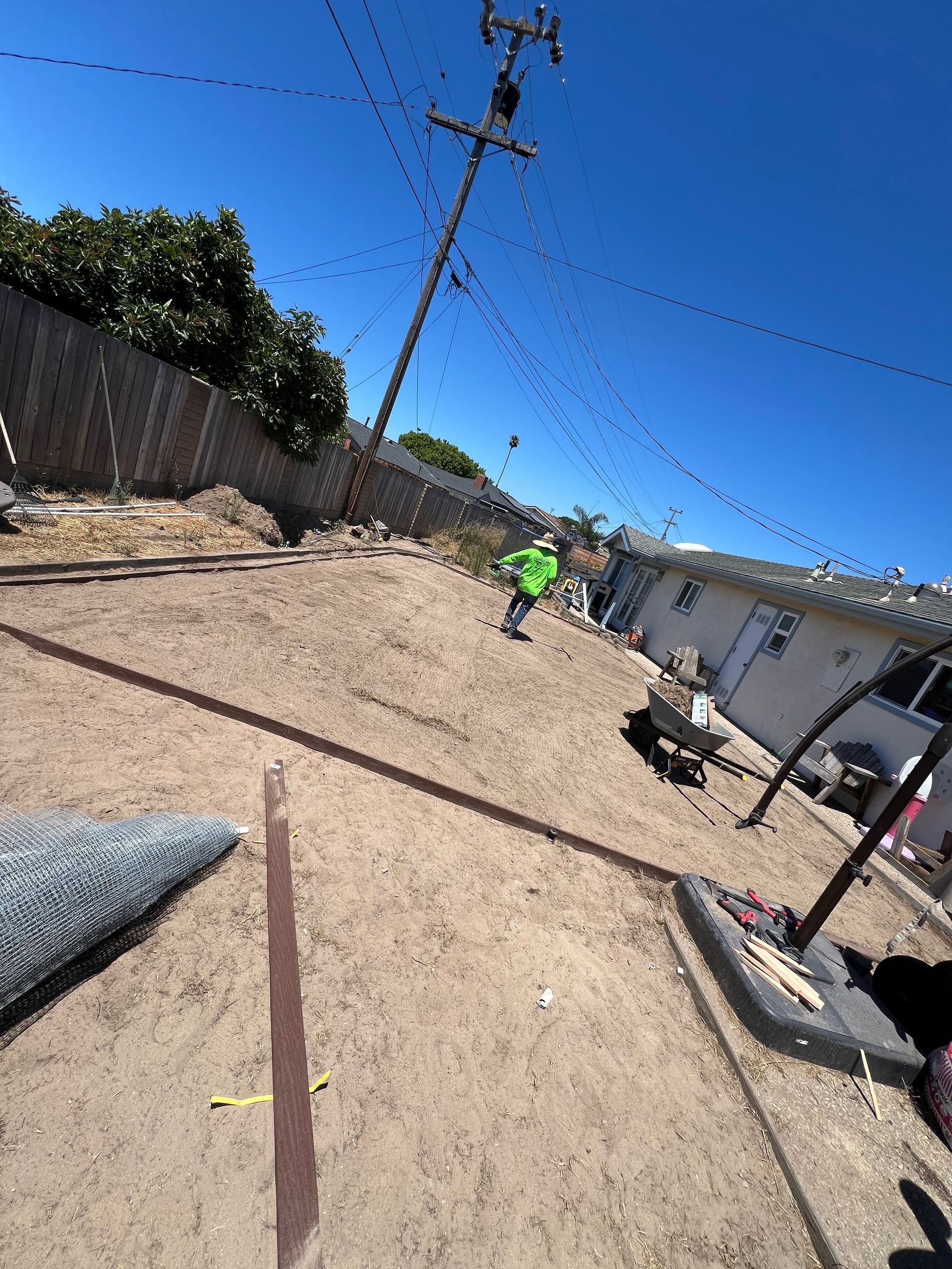 Person in green shirt in a dirt yard, next to a utility pole and fence, under a blue sky.