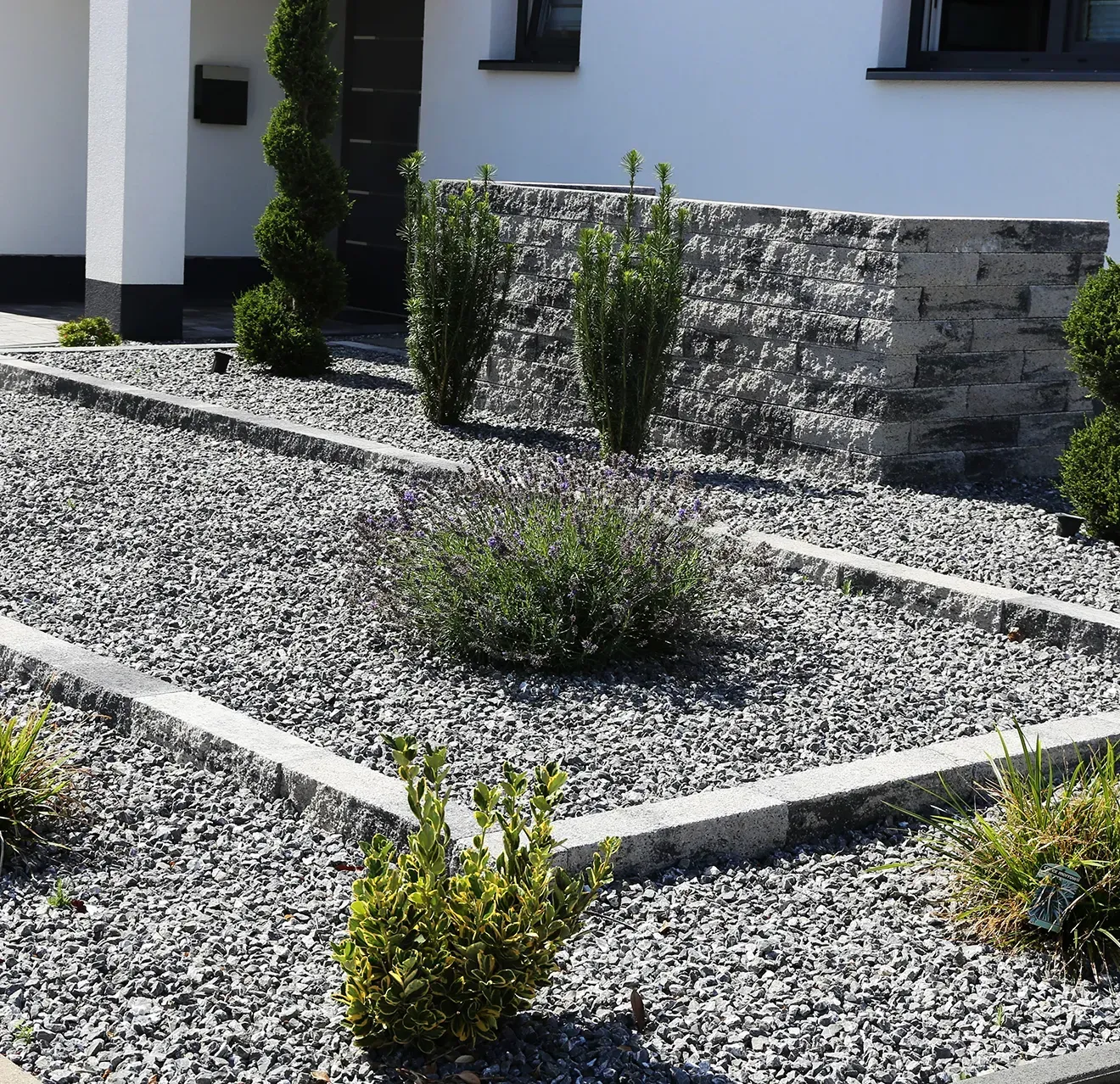 Front yard with a stone retaining wall, gray gravel, and various green shrubs.