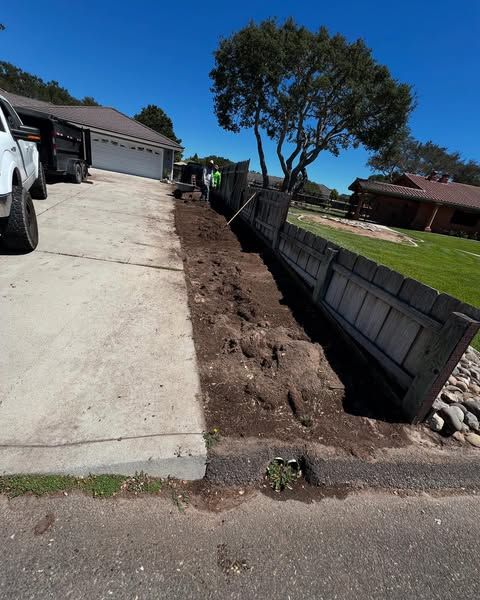 Workers removing a damaged wooden fence next to a driveway and road, under a sunny sky.