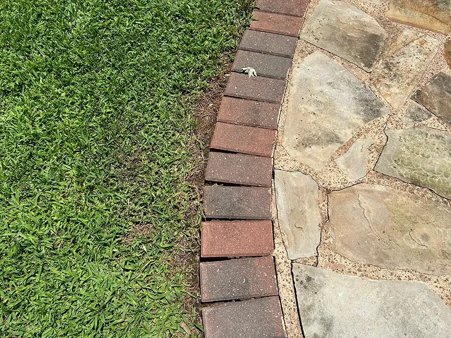 Person using a string trimmer to edge a brick walkway, wearing green boots.