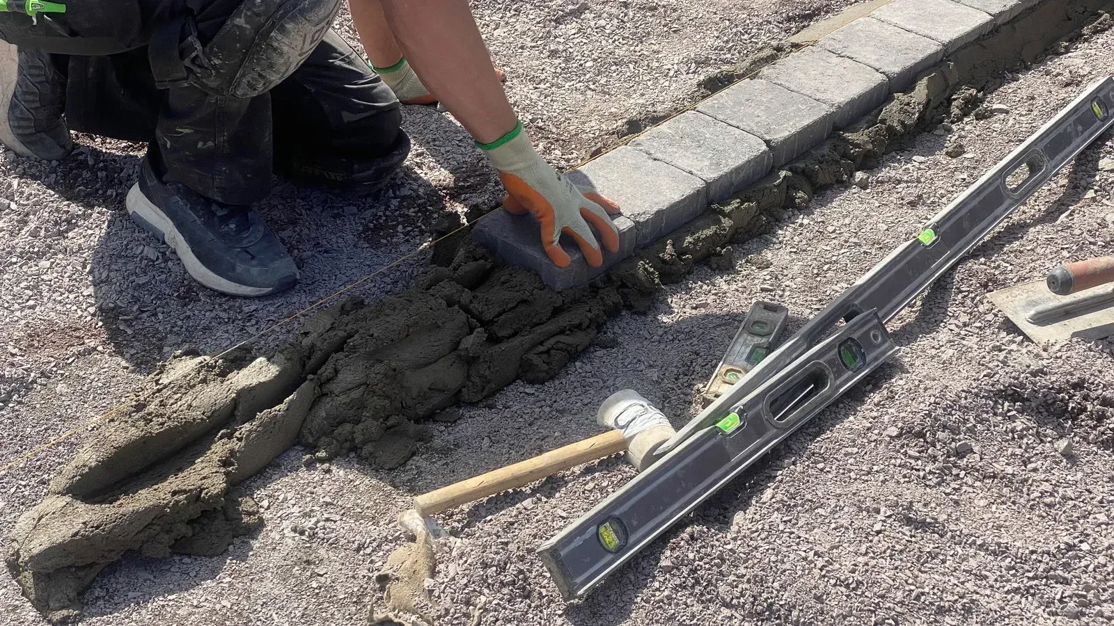 Hands in green gloves hammering edging into soil, gravel on the left, grass on the right.