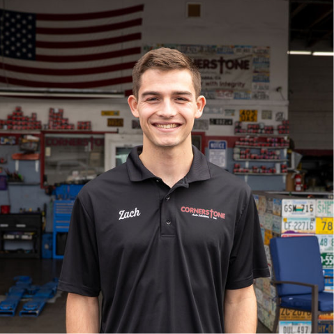 A smiling employee named Zach stands in a service shop wearing a black collared polo with a Cornerstone logo. | Cornerstone Auto Solutions
