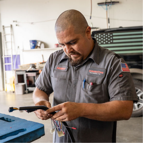 A technician in a grey uniform focuses on wiring and electrical components inside an automotive repair shop. | Cornerstone Auto Solutions