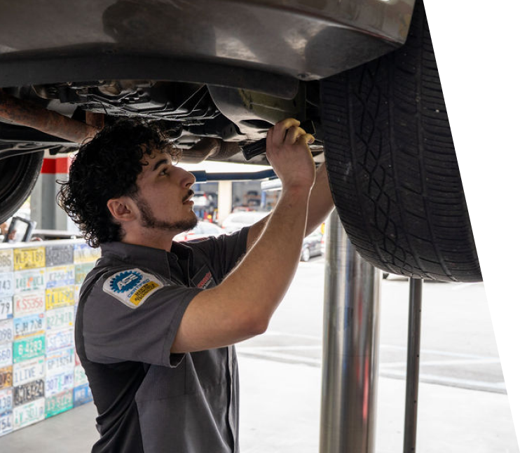A mechanic in a grey uniform inspects the undercarriage of a car raised on a lift in a shop with license plates on the wall. | Cornerstone Auto Solutions