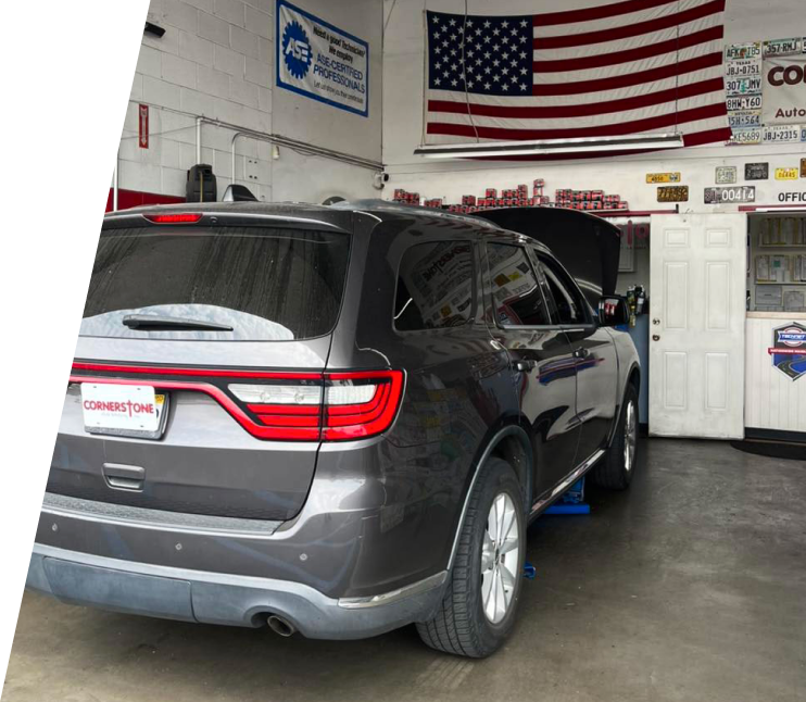 A dark gray SUV parked inside an auto repair shop with the hood raised, beneath a large American flag on the wall. | Cornerstone Auto Solutions