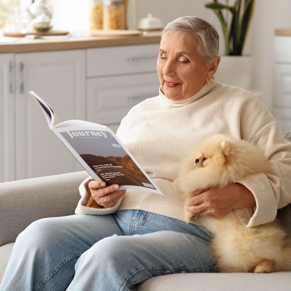 A woman is sitting on a couch holding a dog and reading a magazine.