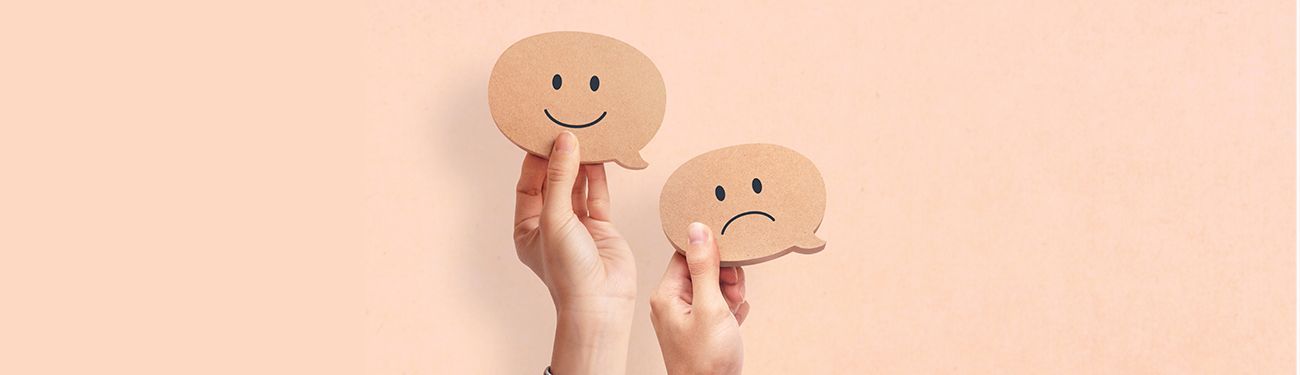 Hands holding two cardboard speech bubbles, one with a happy face, the other with a sad face.