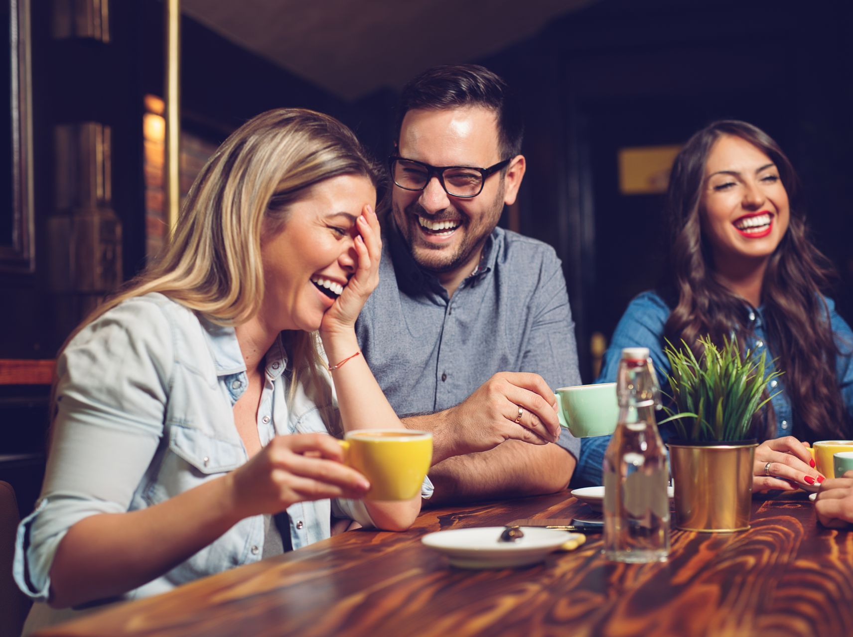 Three people laughing at a table with coffee cups. Interior setting, warm colors.