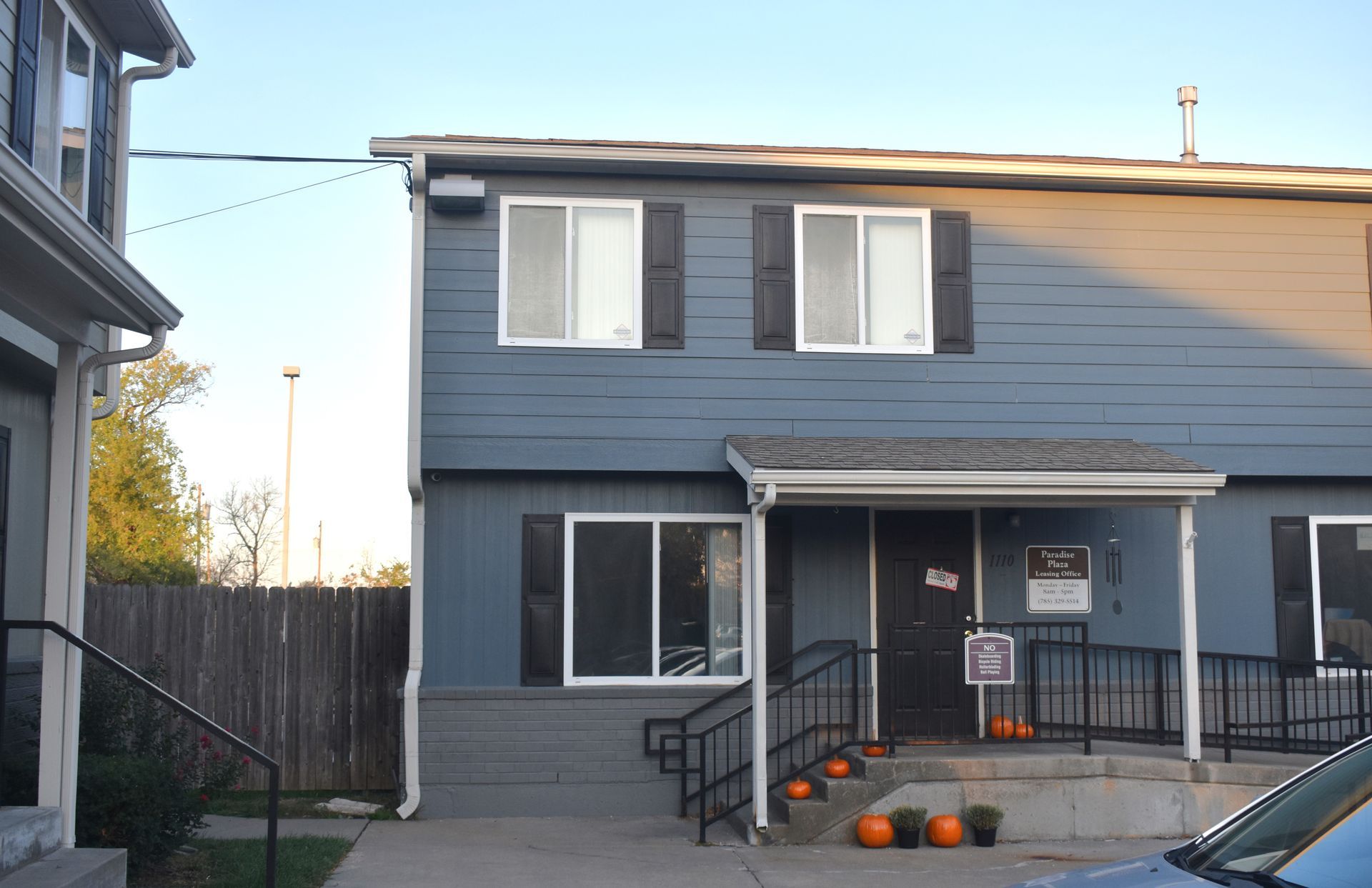 Two-story blue building with white-framed windows, porch, and pumpkins; wooden fence and blue sky in background.