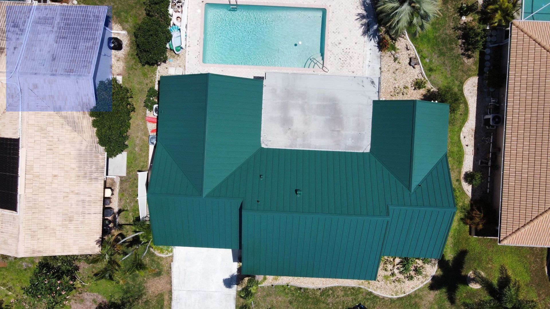 Aerial view of a house with a green roof, a pool, and other houses in a sunny neighborhood.