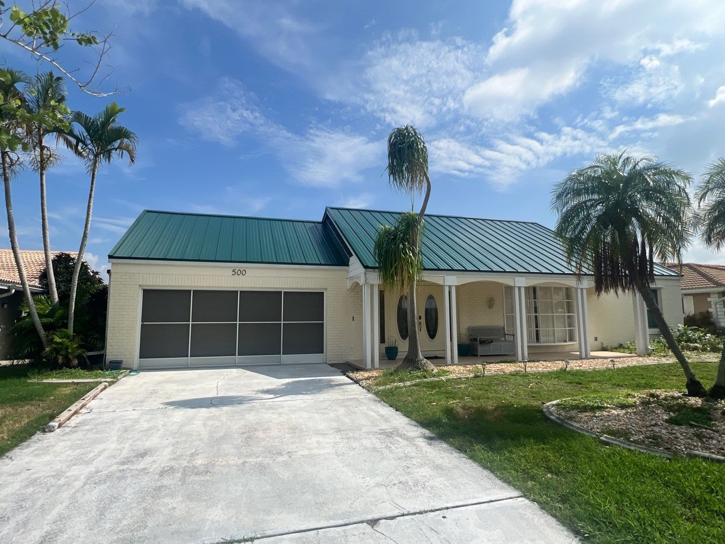 Beige house with green roof, gray garage door, and driveway under a blue sky with palm trees.